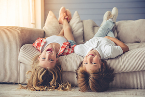 Zwei Geschwister haengen laechelnd kopfueber von der Couch, waehrend die Babysitterin Abenteuer-Geschichten vorliest